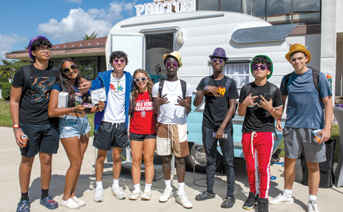 Eight students smiling on campus in front of a photo booth trailer